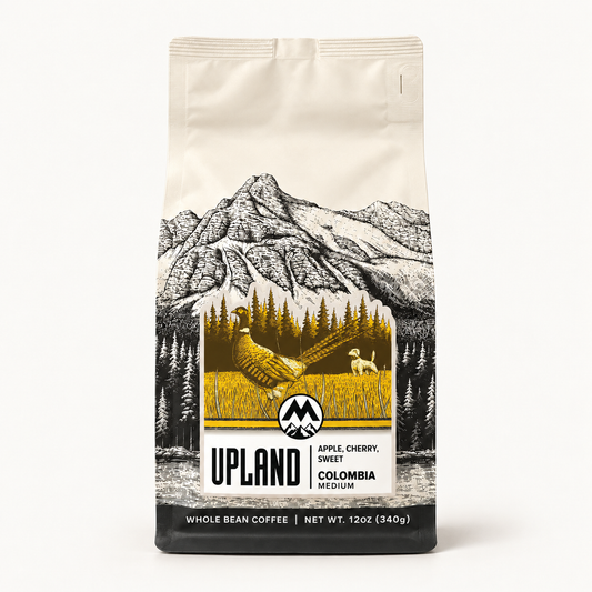 A 12 oz bag of Upland Coffee Company whole bean coffee, Colombia Medium. The packaging features a cream-colored top half with a black line art illustration of a mountain range, and a lower black section with a central golden-yellow graphic of a pheasant in a field alongside a small dog.